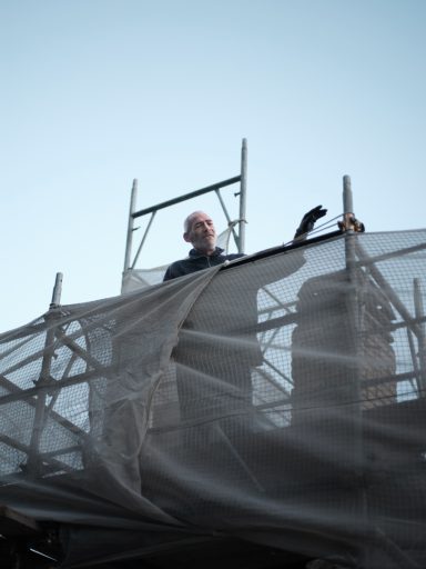 Uomo in cima a una struttura di impalcatura con mano sollevata, coperto da una rete.